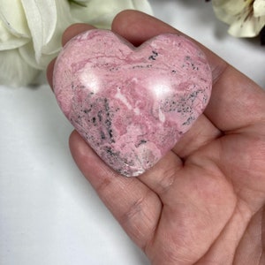 May include: A pink and black heart-shaped polished stone. The stone has a marbled pattern and is smooth to the touch.