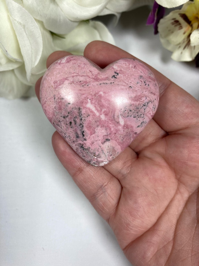 May include: A pink and black heart-shaped polished stone. The stone has a marbled pattern and is smooth to the touch.