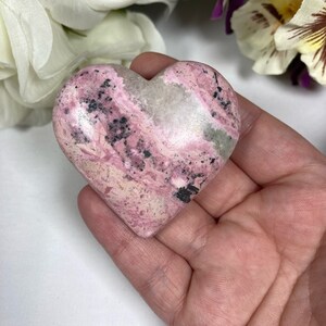 May include: A pink and white heart-shaped stone with black and gray swirls. The stone is smooth and polished.