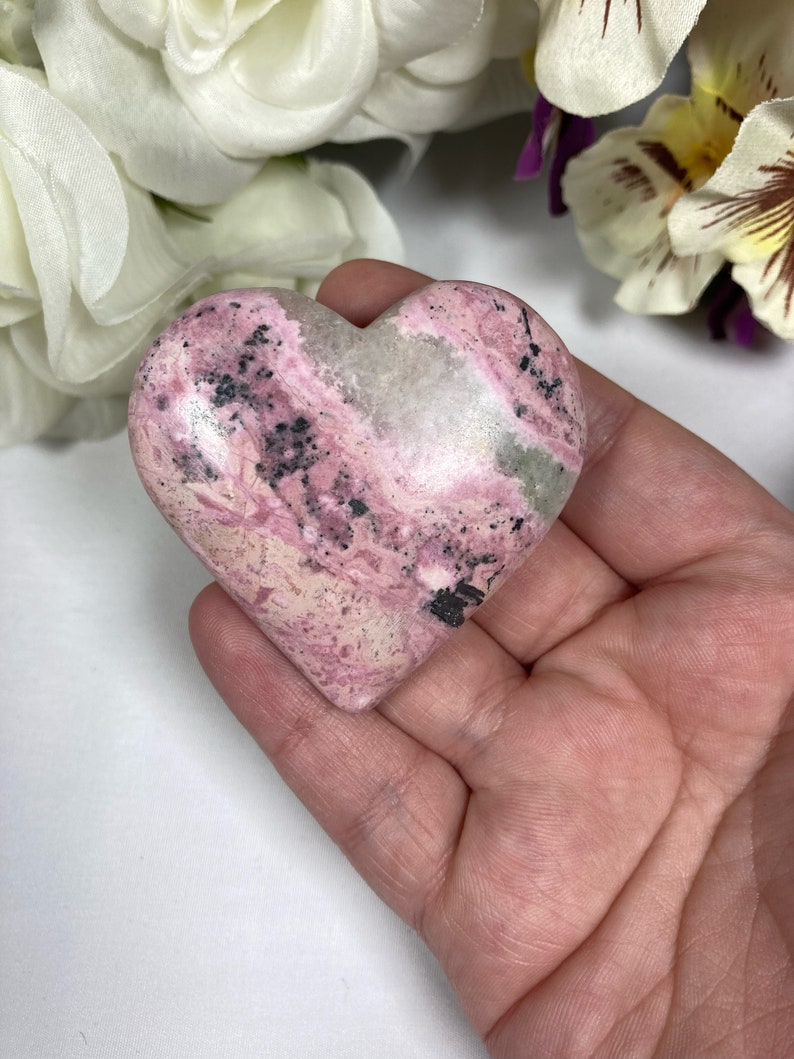 May include: A pink and white heart-shaped stone with black and gray swirls. The stone is smooth and polished.