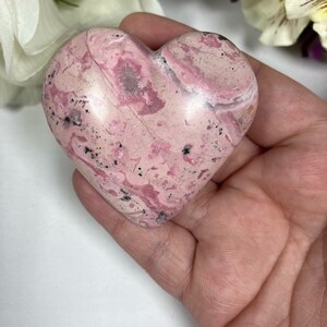 May include: A heart-shaped pink and white polished stone with black flecks. The stone is smooth and has a natural, organic look.