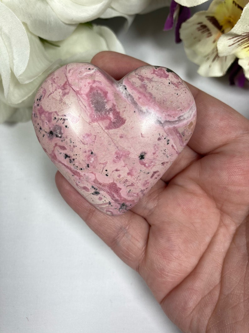 May include: A heart-shaped pink and white polished stone with black flecks. The stone is smooth and has a natural, organic look.