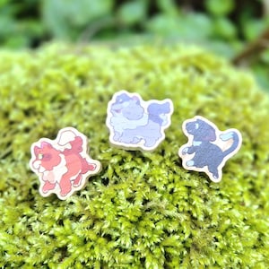 May include: Three wooden cat-shaped pins with cartoon designs. One is red and white, another is light blue and purple, and the third is black and light blue. The pins are set on a bed of green moss.