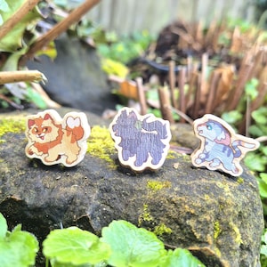 May include: Three wooden animal pins: a ginger cat, a purple dog, and a blue and grey creature. The pins are displayed on a moss-covered rock, with greenery in the background. Each pin is a unique design.