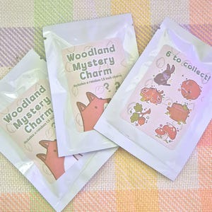 May include: Three sealed white pouches with "Woodland Mystery Charm" labels featuring a fox illustration. One label reads "6 to collect!" with woodland creature graphics. Each charm is 1.5 inches.