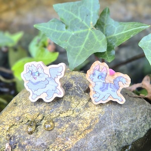 May include: Two wooden cat-shaped earrings. One is light blue with wings, the other is grey with blue stripes. The earrings are set on a rock with green leaves in the background. The earrings are small and detailed.