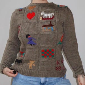 Könnte beinhalten: Ein brauner Pullover mit bunten, gestickten Patches, darunter ein rotes Herz und ein weißes Pferd. Die Patches zeigen auch eine Person in einem lila Kleid und andere Designs. Der Pullover wird mit hellblauen Jeans getragen.