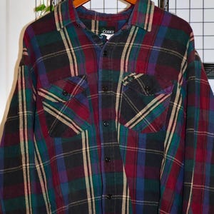 May include: A multi-coloured plaid shirt with long sleeves, featuring a pattern of black, red, green, and purple. The shirt has two chest pockets and a button-down front. The brand name "CODET" is visible on the collar.