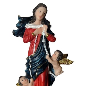 May include: A figurine of the Virgin Mary, known as the Virgin of the Untied Knots, depicted in a red dress and blue robe, holding a rope in her hands. Two cherubs are flanking her, and a snake is at her feet. The base of the figurine is black with gold accents and the text "Virgen Desatanudos" is inscribed on it.