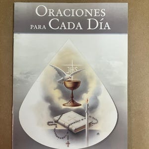 May include: A book cover with the title "Oraciones Para Cada Día" in white letters. The cover features a dove, chalice, book, rosary, and candle within a white teardrop shape against a gray background.