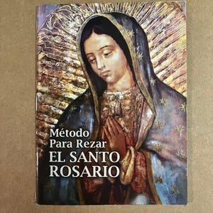 May include: A book titled "Método Para Rezar El Santo Rosario" featuring an illustration of a woman with a blue veil and clasped hands. The cover has gold and brown tones with text in white.