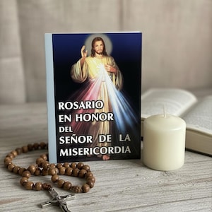 May include: A book titled "ROSARIO EN HONOR DEL SEÑOR DE LA MISERICORDIA" with an image of Jesus Christ, a rosary, a white candle, and an open book on a wooden surface. The rosary has brown beads and a silver cross.