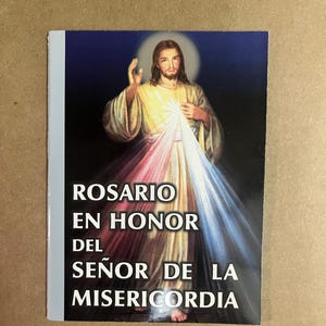 May include: A religious booklet with the title "ROSARIO EN HONOR DEL SEÑOR DE LA MISERICORDIA" in white text. The cover features an illustration of Jesus with a halo, radiating light in shades of yellow, white, and red. The background is dark blue.