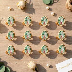 May include: Gold-toned rings with oval, painted inserts featuring an angel design. The rings are arranged on a light wooden surface, accompanied by pearls, dried roses, and a tag that reads "Handcrafted Blessings."