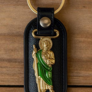 May include: Black leather keychain with a gold-colored metal Saint Jude figure, wearing a green robe. The keychain has a gold-colored ring and a gold-colored metal clasp. The background is a wooden surface.