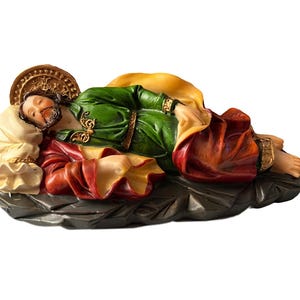 May include: A figurine of a sleeping saint, lying on a bed of rock. The saint is wearing a green robe with gold trim, a red robe, and a gold halo. The figurine is made of painted resin.