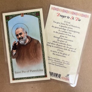 May include: Two prayer cards in protective sleeves. One card features an image of Saint Pio of Pietrelcina with text below. The other card has a prayer to Saint Pio printed on it. The cards are on a light brown surface.