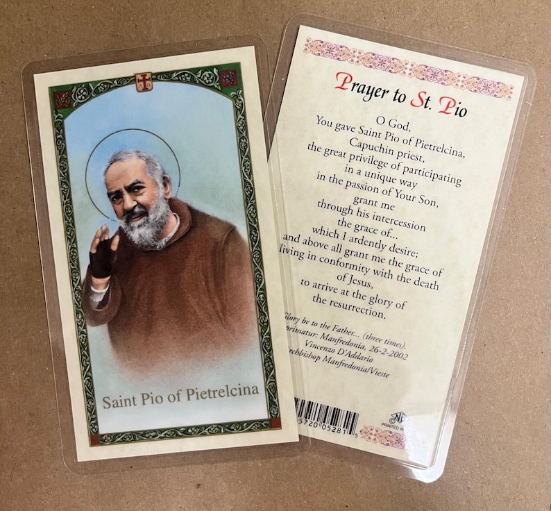 May include: Two prayer cards in protective sleeves. One card features an image of Saint Pio of Pietrelcina with text below. The other card has a prayer to Saint Pio printed on it. The cards are on a light brown surface.