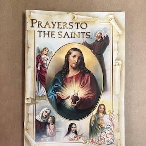 May include: A religious book titled "Prayers to the Saints" with illustrations of religious figures. The cover features a central image of Jesus holding a glowing heart, surrounded by other saints and angels. The book has a vintage, parchment-like design.