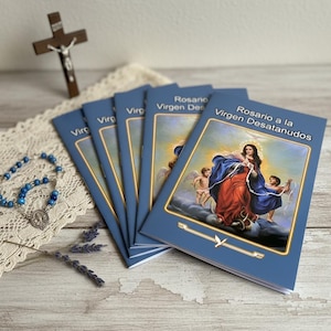 May include: A stack of blue booklets titled "Rosario a la Virgen Desatanudos" with an image of the Virgin Mary. A rosary with blue beads and a wooden crucifix are also visible. The items are arranged on a white lace cloth.