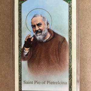 May include: A religious card featuring an illustration of Saint Pio of Pietrelcina. The image shows a man with a white beard and a halo, wearing a brown robe. The card has a decorative border and the saint's name is printed at the bottom.