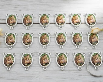 May include: Oval-shaped religious charms with a silver-tone metal frame. Each charm features a painted image of a saint holding a child, surrounded by a green border. The charms are arranged on a light wood-grain surface.