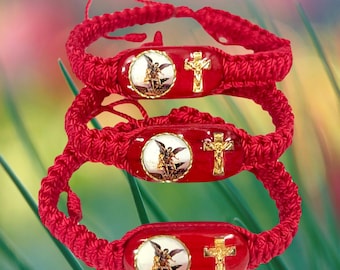 May include: Three red braided bracelets with a gold cross and an image of an angel on each. The bracelets are stacked on top of each other.