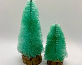 May include: Two green bottle brush Christmas trees on wooden bases. The trees are different sizes.
