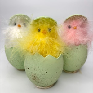 May include: Three pastel colored chicks peeking out of cracked green eggs. The chicks are yellow, white, and pink with fluffy feathers.