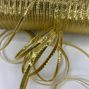 May include: A spool of gold metallic ribbon with a textured surface. The ribbon is wound around the spool and is ready to be used for crafting or decorating.