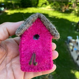 May include: A small pink felt birdhouse with a brown felt roof and a small hole for the birds to enter. The birdhouse is held in a hand.