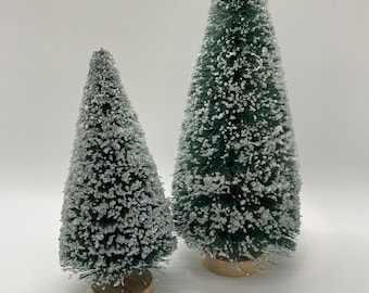 May include: Two artificial Christmas trees. The trees are green with white, snow-like accents. They are cone-shaped and sit on small, round, wooden bases. The trees vary in size, with one being taller than the other.