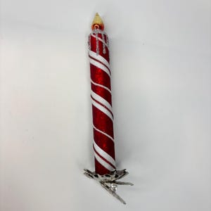 May include: A red and white striped candle ornament with a silver clip base. The candle has a glittery finish and a yellow flame tip. The ornament is designed for holiday decor.