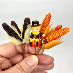 May include: Thanksgiving-themed decorative picks featuring miniature corn cobs and a pilgrim figure. The corn cobs are in shades of orange and dark purple, with a small pilgrim figurine in the center. The figurine is wearing a black hat and a yellow shirt.