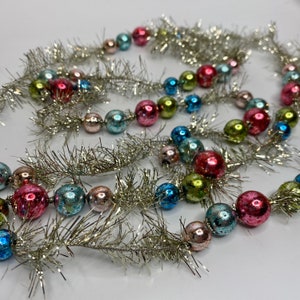 May include: A decorative garland featuring a silver metallic tinsel strand interspersed with small, round, reflective glass beads in various colors including pink, light blue, lime green, and metallic silver, arranged in a repeating pattern.