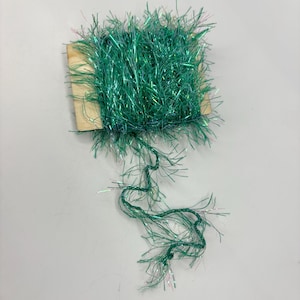 May include: A spool of vibrant green tinsel yarn, tightly wound and secured to a small wooden base. The yarn has a metallic sheen, reflecting light. Strands of the yarn extend from the spool, creating a textured appearance. Ideal for crafting and decorative projects.