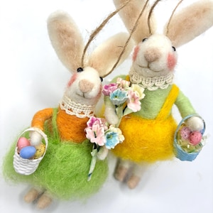 May include: Two white felt bunny ornaments with long ears, holding baskets of colorful Easter eggs and bouquets of pastel flowers. The bunnies are wearing green and yellow dresses with white lace collars.