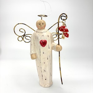 May include: A decorative angel figurine with a distressed white robe, a red heart emblem, and gold-toned wire wings. The figure holds a tall, thin, dark metal staff with two small, glossy red mushroom charms hanging from the top hook.