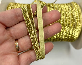 May include: A spool of gold glitter ribbon with a beige elastic band. The ribbon is wrapped around the spool and is held in a hand.