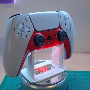 Peut inclure: Une manette de jeu PlayStation 5 blanche et rouge est exposée sur un support en plastique blanc. Le support a une petite fenêtre rectangulaire avec le texte "[CUSTOM TEXT]" imprimé dessus.