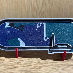 以下が含まれることがあります： 青と緑の表面を持つレーシングトラックの3Dプリントモデル。トラックは赤い支えのある白いスタンドの上にあります。