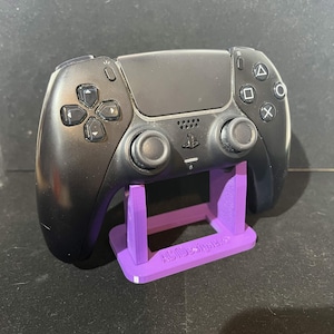 May include: A black video game controller rests on a purple stand. The controller has buttons and joysticks. The stand is rectangular with the text "ABIDesigners" on the side.
