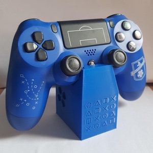 May include: A blue PlayStation 4 controller with a blue charging stand. The controller has a soccer field design on the touchpad and a soccer ball logo on the back. The stand has the PlayStation button symbols on the front.