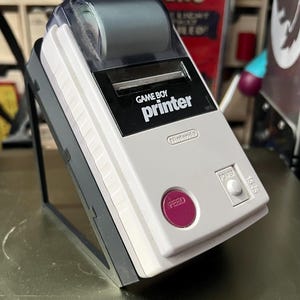 Op de afbeelding: Een witte Game Boy Printer met een zwart en grijs ontwerp. De printer heeft een rol thermisch papier erin en een paarse knop met het label "Feed". De printer staat op een zwarte standaard.
