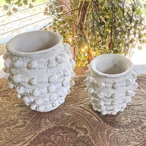 May include: Two off-white ceramic vessels with a textured, bumpy surface featuring raised circular nodes arranged in horizontal bands. Both containers have a wide, open top and a rounded, bulbous shape, displaying a matte, weathered finish.