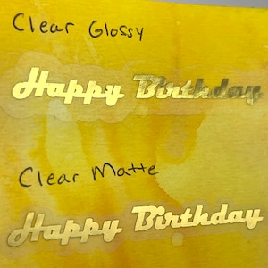 May include: Two clear stickers with gold lettering that reads "Happy Birthday". One sticker is labeled "Clear Glossy" and the other is labeled "Clear Matte". The stickers are on a yellow and orange watercolor background.