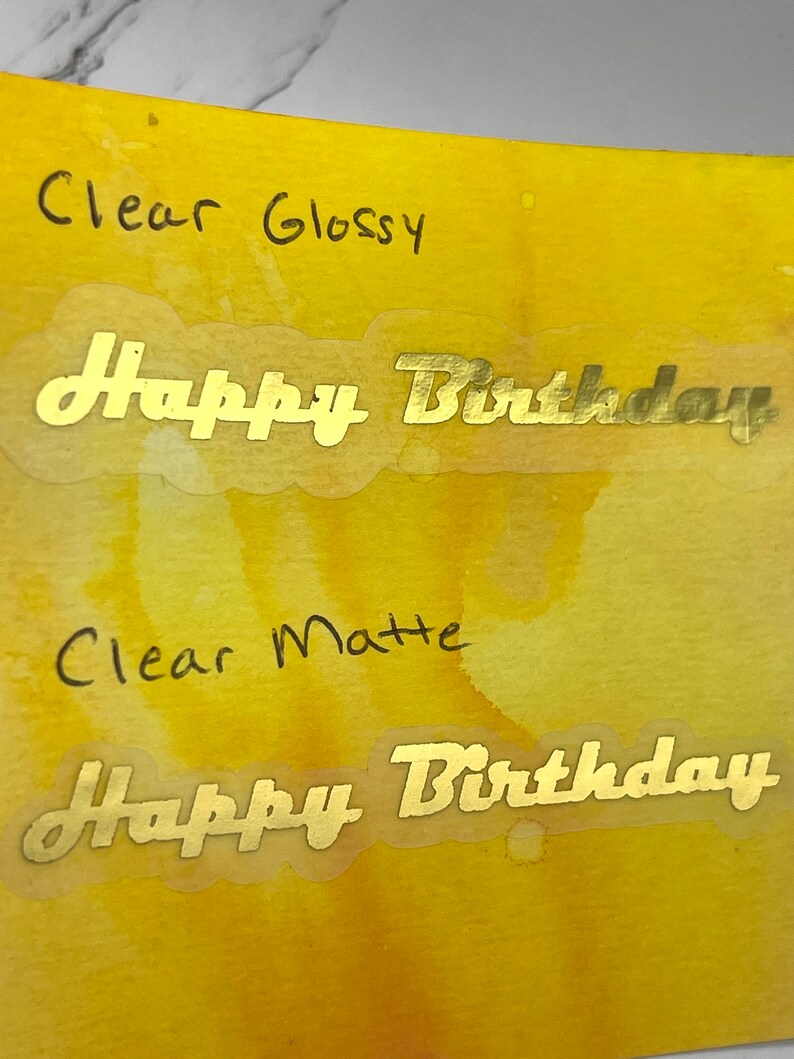 May include: Two clear stickers with gold lettering that reads "Happy Birthday". One sticker is labeled "Clear Glossy" and the other is labeled "Clear Matte". The stickers are on a yellow and orange watercolor background.