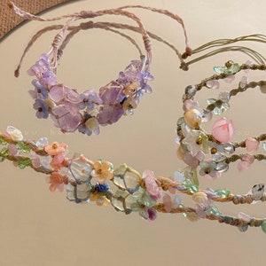 May include: Three bracelets made with colourful stones and flowers. The bracelets are made with a variety of stones, including amethyst, rose quartz, and green aventurine. The flowers are made of white and blue beads. The bracelets are tied with a brown cord.