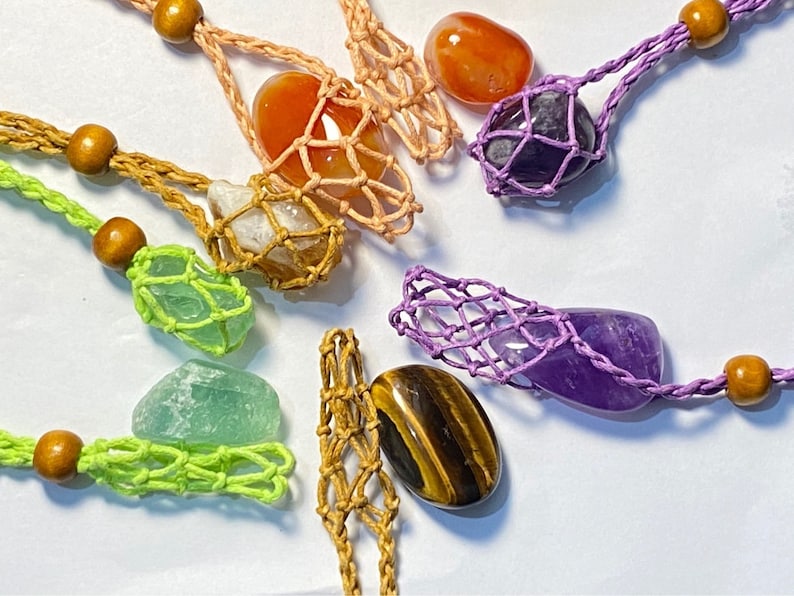 May include: A collection of seven colorful crystal stones, each encased in a crocheted net bag. The stones include amethyst, carnelian, tiger's eye, clear quartz, green aventurine, and two other stones.