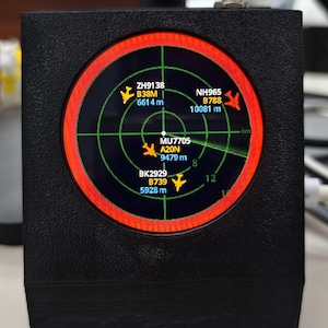 May include: A black, textured, wedge-shaped electronic device featuring a circular digital radar display. The screen shows green grid lines, a red circular border, and yellow airplane icons with text labels including ZH9138, NH965, MU7705, and BK2929.
